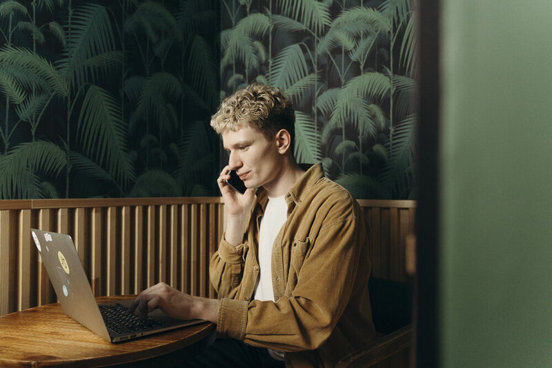 This image shows the profile of a white masculine-presenting person with curly short blond hair, holding a phone to their ear, opposite hand resting on a laptop keyboard. They are wearing a tan corduroy button-down, and a dark green tropical wallpaper covers the wall behind them.