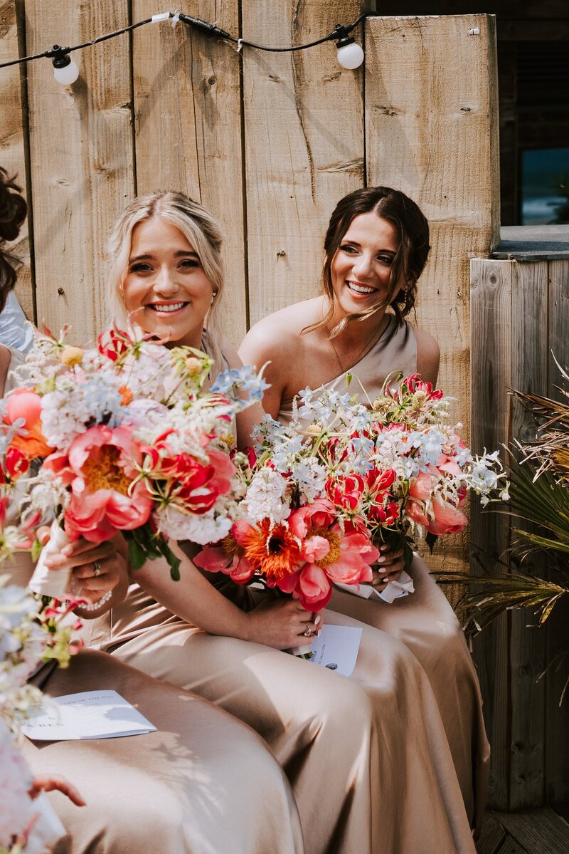 Bridesmaids in oyster coloured dresses holding bouquets of coral and blue flowers, including coral charm peonies and blue delphiniums