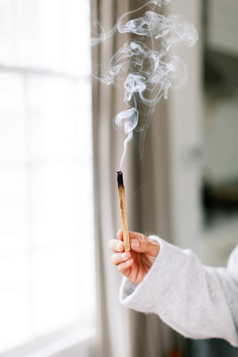 A lit incense stick with smoke curling upwards, against a blurred background.