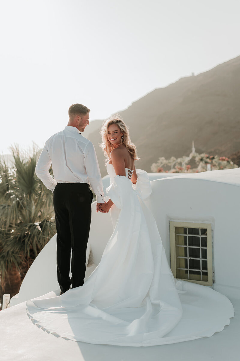 Bride and Groom smile holding hands on the white stone rooftop in Santorini, Greece