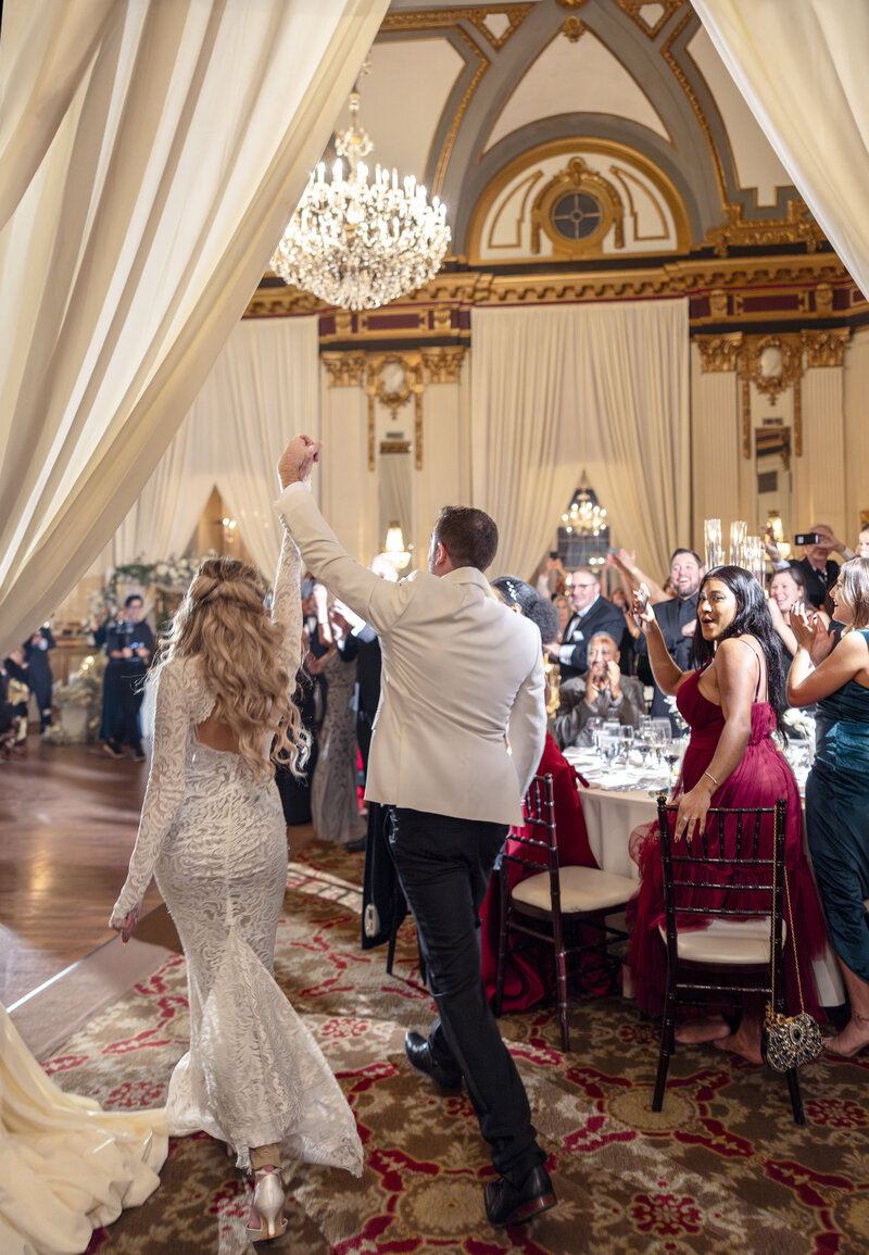 A couple in formal attire, with the woman in a white gown and the man in a white jacket, joyfully enters a lavish ballroom with guests applauding. The room is elegantly decorated with chandeliers, draped curtains, and richly detailed ceilings.