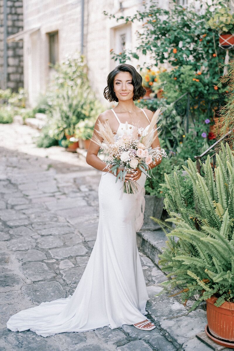 bride-with-bouquet-stands-near-stone-house-with-many-flower-pots