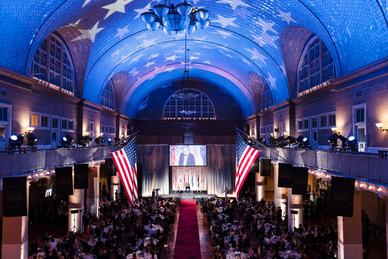 Large NYC corporate gala photographed inside a grand hall with patriotic lighting, banquet tables, and a speaker on stage.