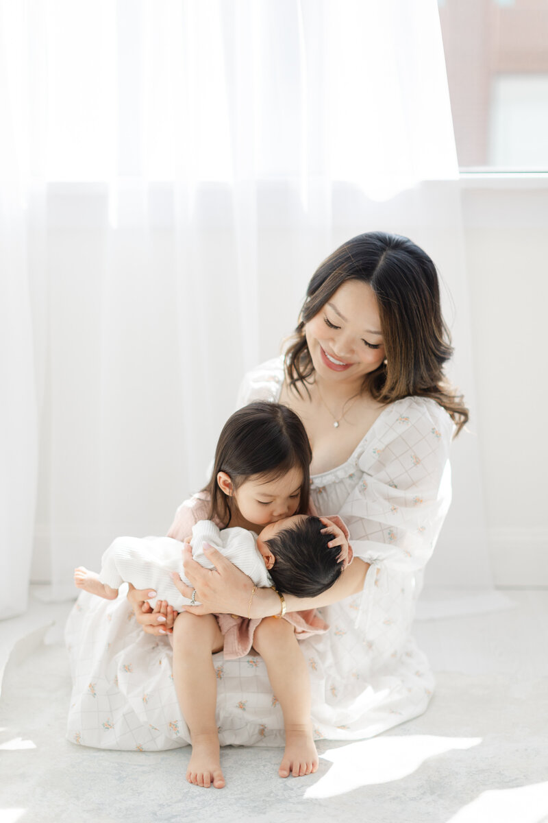 Mom holds young daughter who kisses her newborn baby sibling