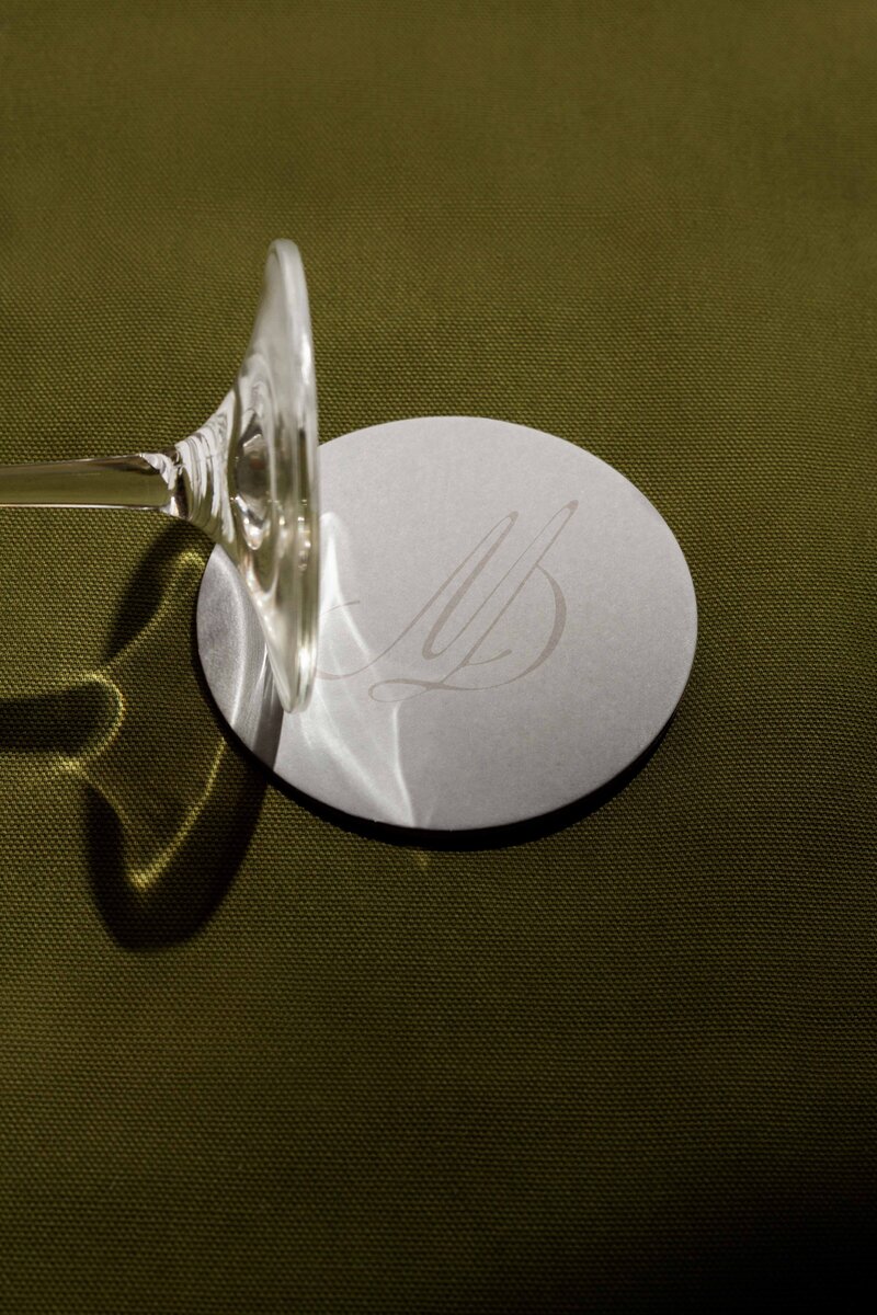 A wine glass rests upside down on a round white coaster with subtle embossed initials on a green textured surface, capturing the refined elegance often celebrated in showit web design and branding.