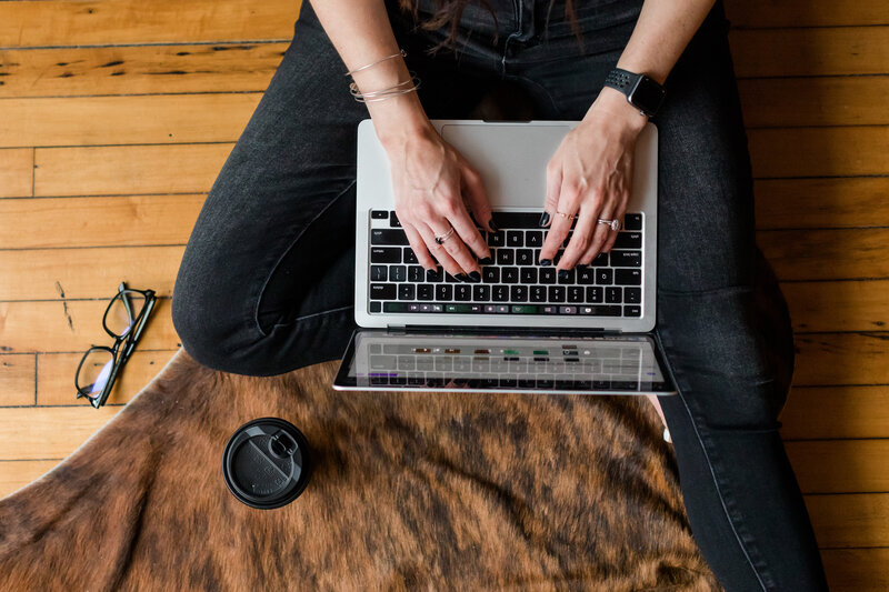 Overhead view of Allie Clark working on her MacBook Pro while seated cross-legged on a patterned rug, actively engaged in the website customization process behind Allie Clark Designs.