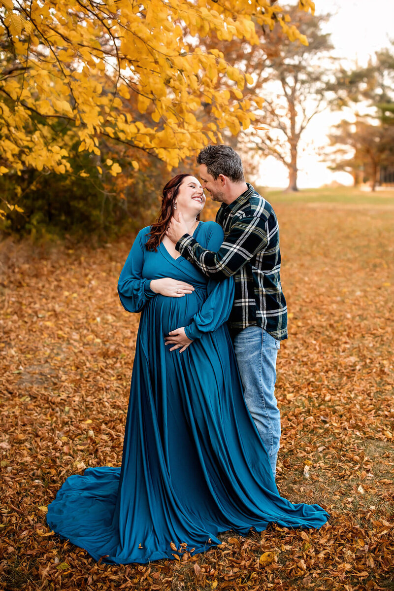 Pregnant woman is embraced by man in plaid shirt while standing in fall foilage.