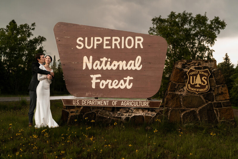 Bride and groom kiss in front of Superior National Forest sign.