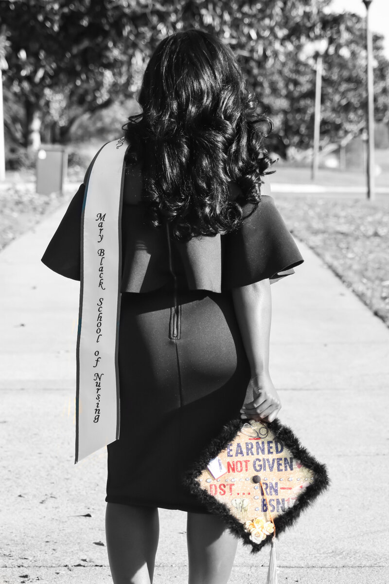 black and white photo of a college graduate with her back to the camera. shes carrying her stole and a graduation cap that reads "earned not given"
