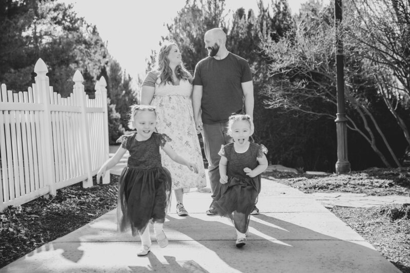 Idaho Falls maternity photos with mother and father holding hands while walking down the sidewalk together and their two daughters run towards the camera
