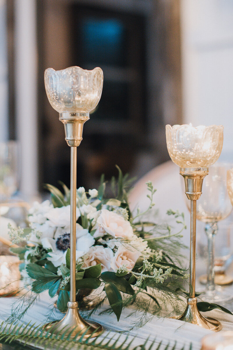 Meercury Glasss flooating candles on brass taper candlesticks are part of a reception table arrangement featuring anemones and roses