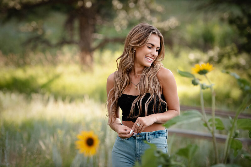 A high school senior giggles down her shoulder while exploring sunflowers in a garden in a black shirt and jeans