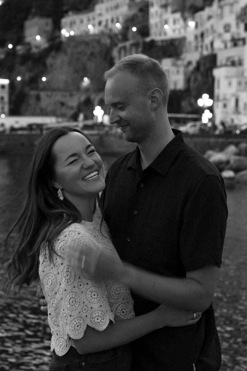 Man and woman smiling and laughing together standing in front of Positano on the Mediterranean sea the night before their wedding on the Amalfi Coast in Italy, by Andrea-Marie Photography