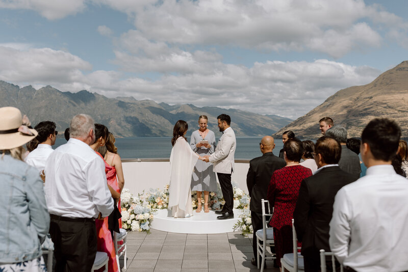 Outdoor wedding ceremony with a couple exchanging vows on a circular platform, surrounded by guests and framed by a stunning lake and mountain backdrop under a partly cloudy sky.