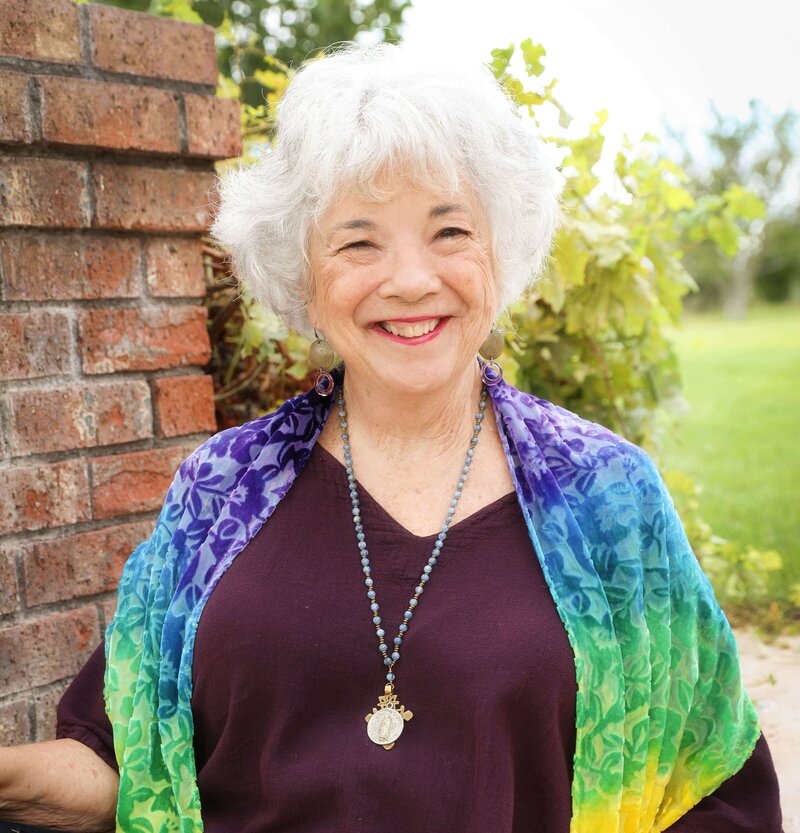Dr. Samarah Grace wearing a purple blouse and rainbow shaw looking at the camera and smiling .  Green field behind her.
