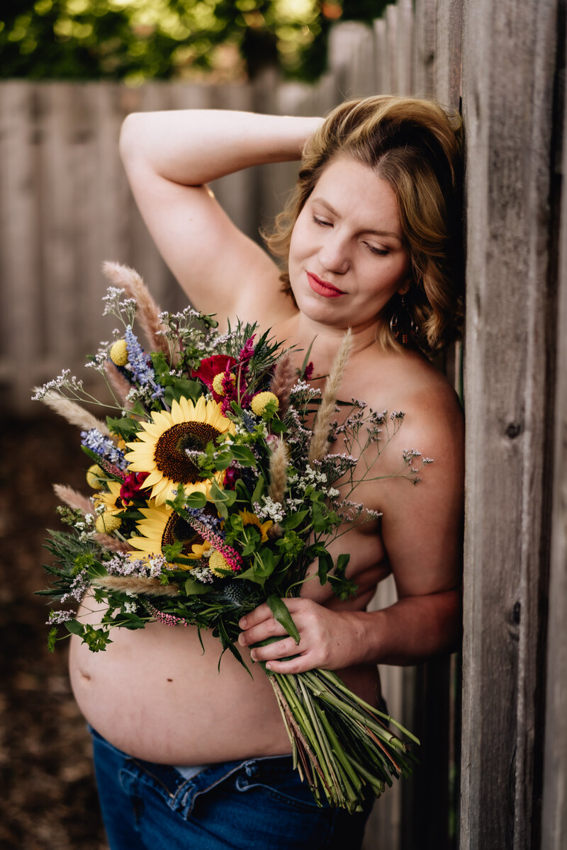 A pregnant woman standing topless in a rustic outdoor setting, holding a bouquet of sunflowers and wildflowers, with one arm resting on a wooden fence. She gazes downwards.