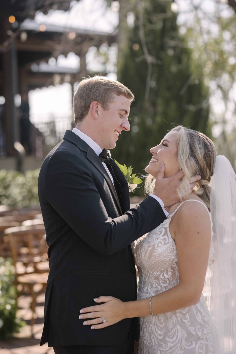 A tender moment between a bride and groom during their black-tie wedding in St. Augustine. The groom, dressed in a sharp black tuxedo, gently holds the bride’s face as they exchange an emotional glance. The bride wears a fitted lace gown and flowing veil, with golden sunlight casting a warm glow over the outdoor ceremony setting. Captured by Phavy Photography in a true-to-color, editorial style that highlights genuine connection, refined details, and timeless elegance.