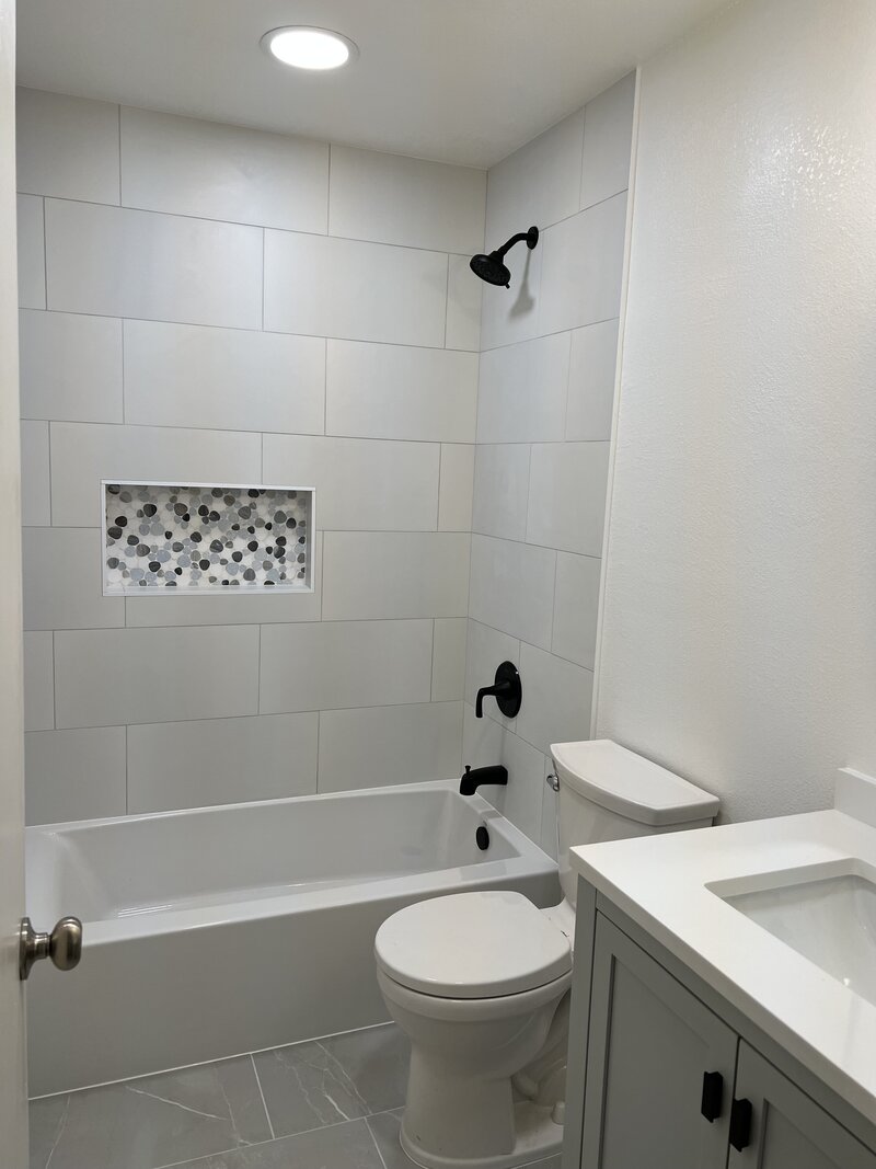A shower remodel with large white tiles placed horizontally. There is a shampoo box with black and white stone pattern and black hardware throughout.