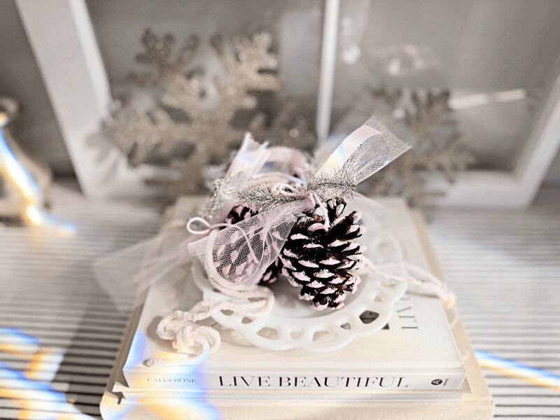 A small white dish holding hand-painted pinecone ornaments with silver ribbon, styled like a holiday ornament, sits atop stacked books titled "Live Beautiful," with silver snowflake decorations in the background—a charming touch by Sweet Pea Ink Creative.