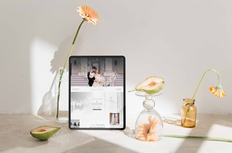 A tablet displaying a wedding photo of a couple is placed on a woven chair next to a notebook under dappled light, captured by talented Showit designers.