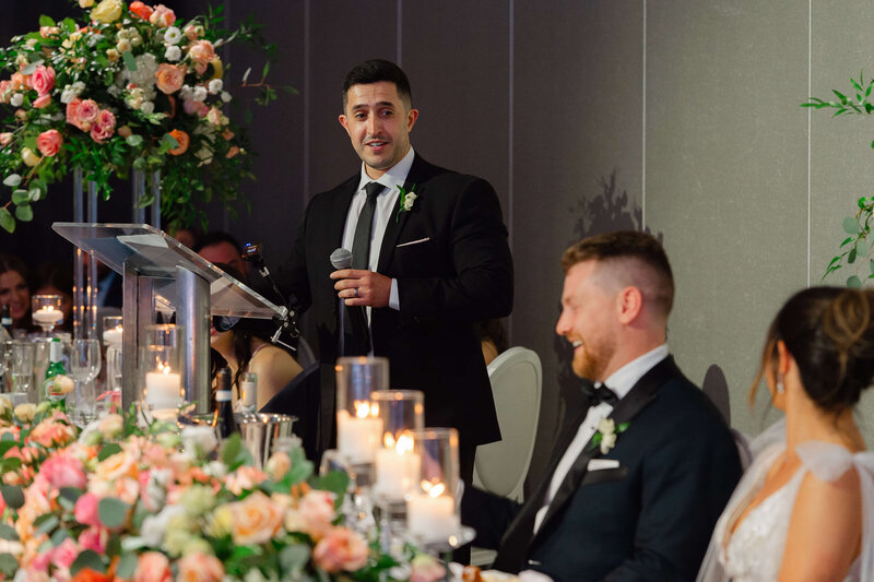 a bride and groom laugh as their best man toasts them.  Captured by Ottawa wedding photographer JEMMAN Photography