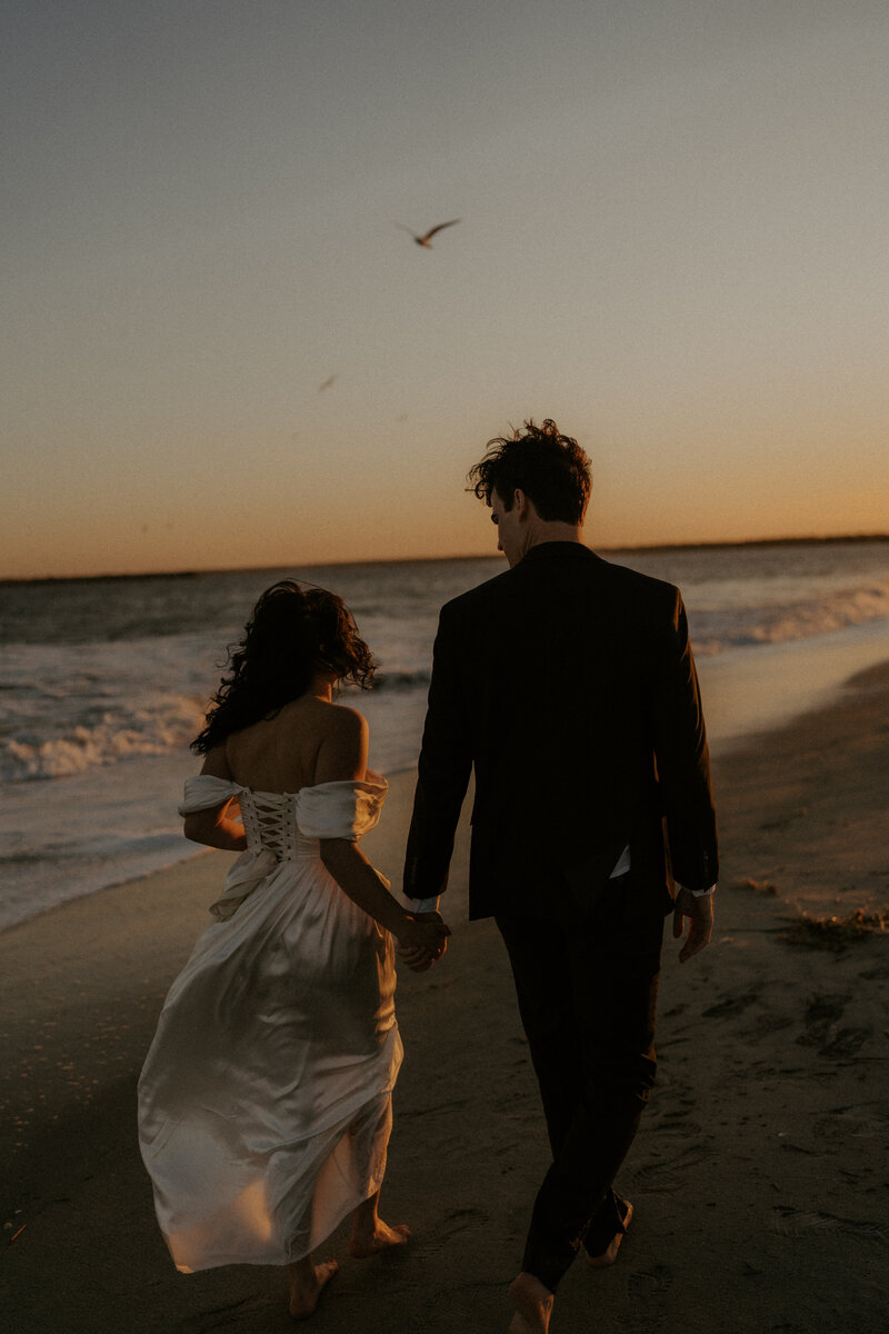 Couple walking hand in hand along the beach, with ocean waves crashing behind them and the sun casting a golden glow.