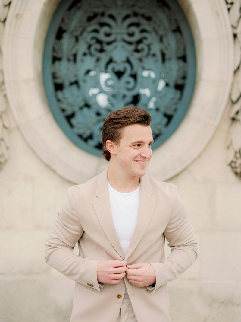 a man with dark slicked back hair looking off into the distance and buttoning up his tan suit jacket in Paris with a french styled building behind him