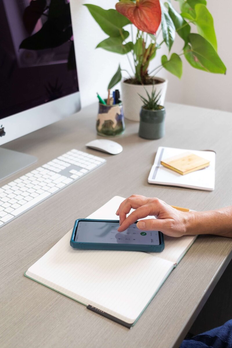 hand touching cell phone on top of desk with computer and plant