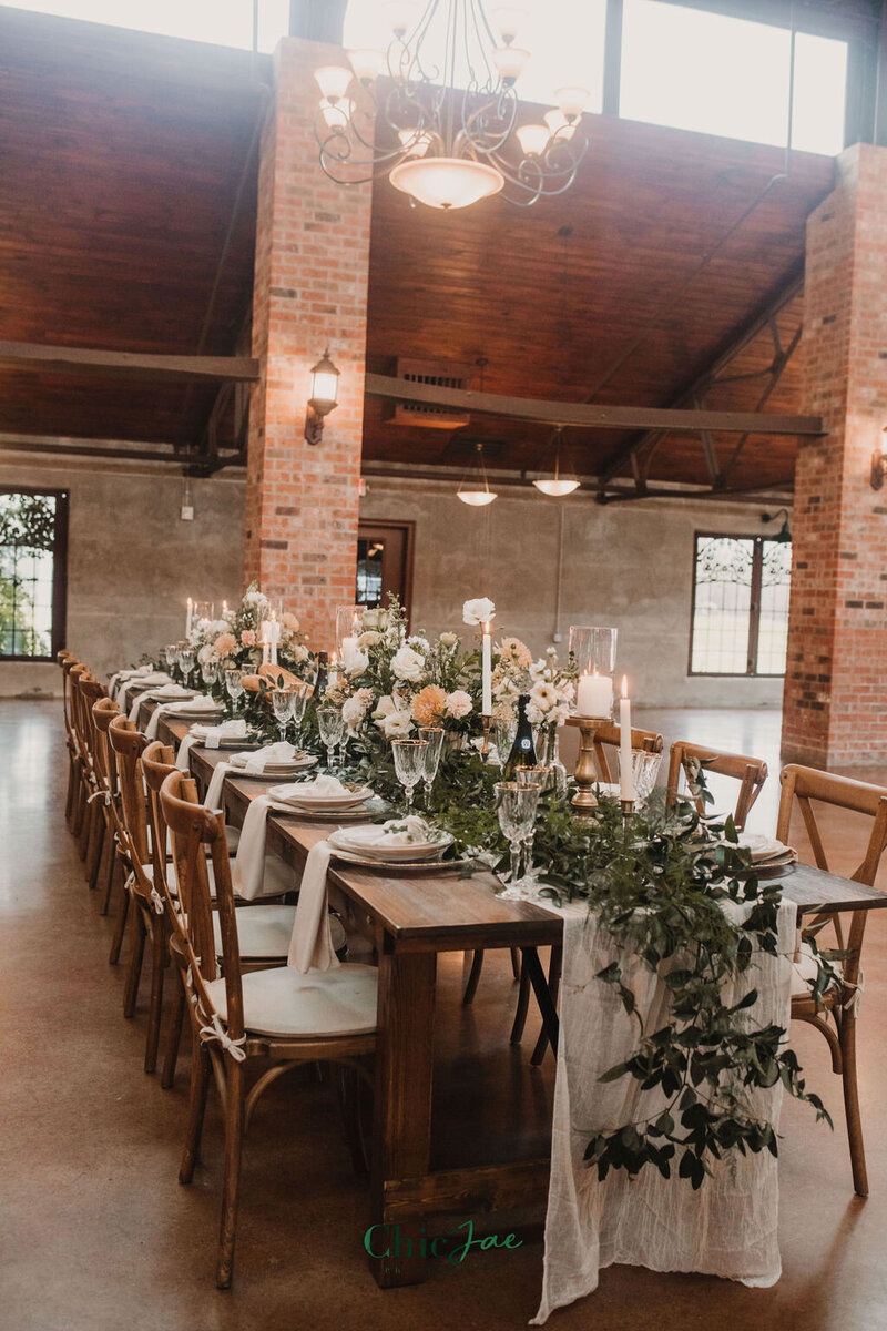 Rustic wedding table with Crossback chairs, ivory runners, green garlands, and taper candles in an open loft-style venue.