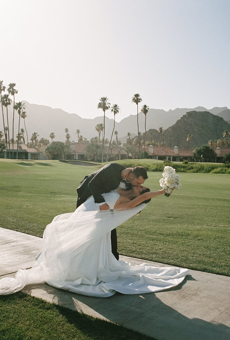 Groom dipping bride as he kisses her and she holds her bouquet in the air with palm tree and mountain landscape in the background