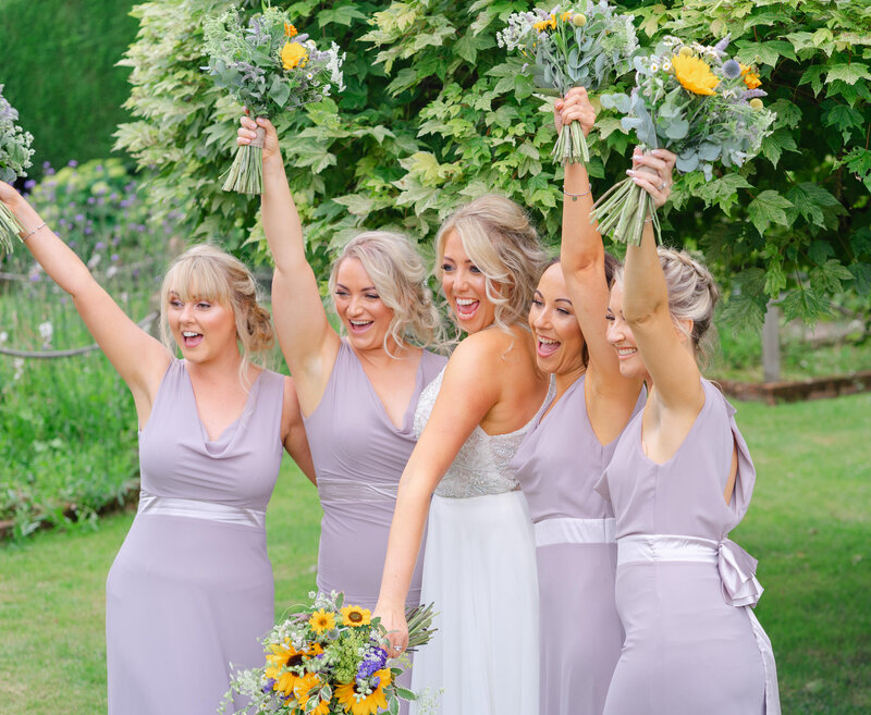 Bridesmaids in matching lilac dresses celebrating with bouquets at a summer wedding in the UK