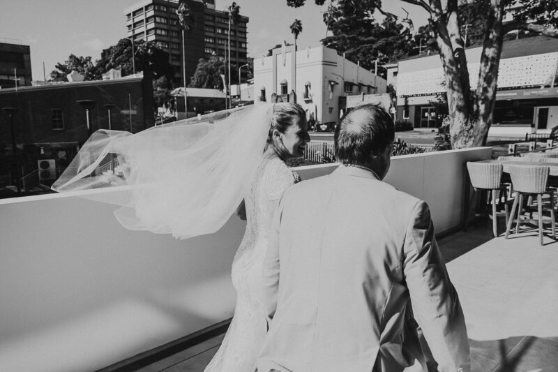 City Hall in Gosford provides a Vegas-style Elopement Experience on the Central Coast. The image shows a couple walking through the city streets with the venue in the background. It is black and white. The bride's veil flow behind her in the wind. It is shot documentary style by Underatreehouse Photography and looks happy and fun