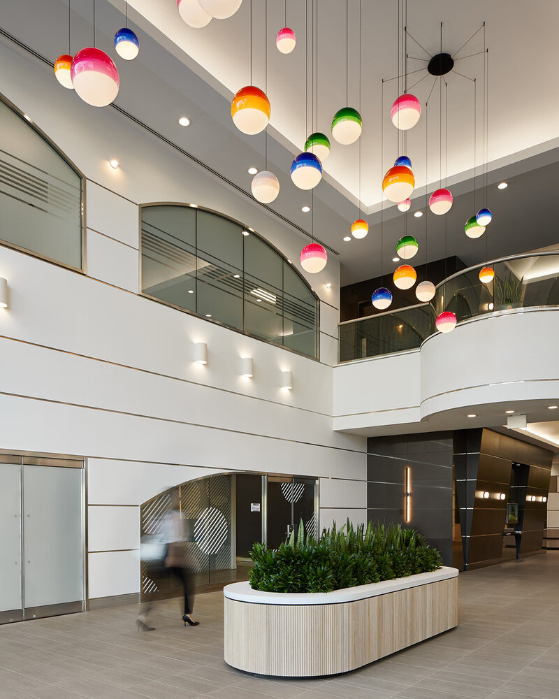 A bright, airy lobby featuring soaring white walls, neutral floor tiles, an indoor planter, a balcony, and a cluster of tritone globe pendant lighting fixtures hanging from the ceiling