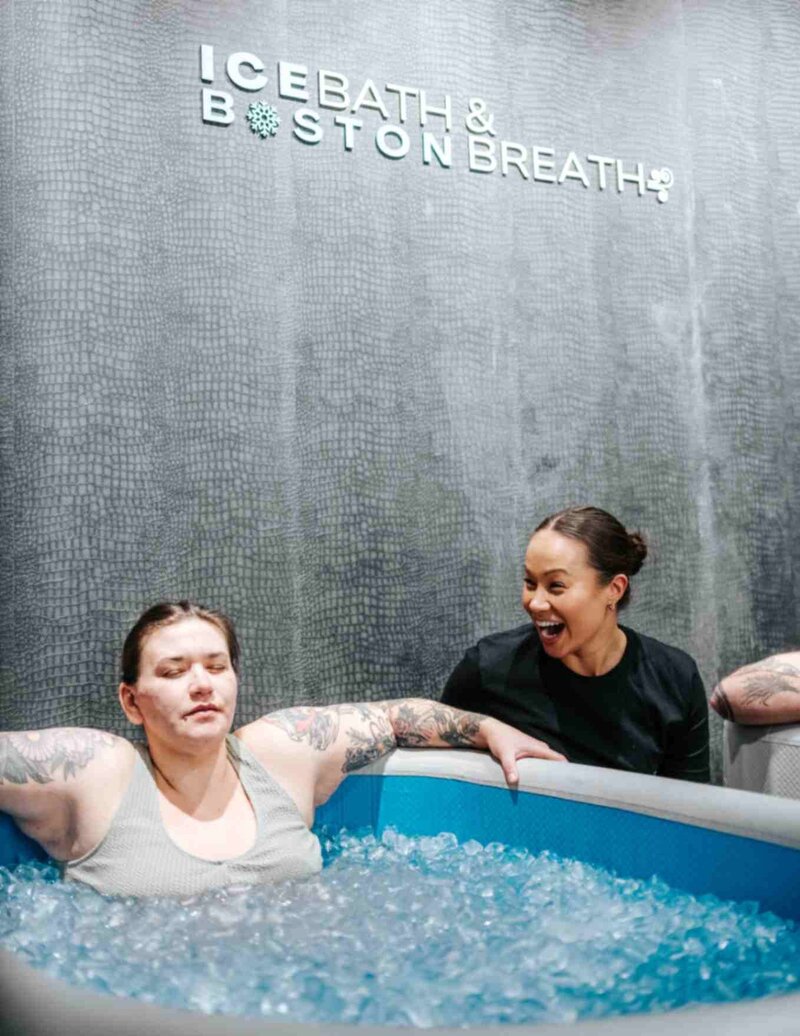 A young tattooed woman smiles breathes deeply and calmly while being guiding through an ice bath as Rachel Lee kneels beside her smiling wide as she provides instruction.