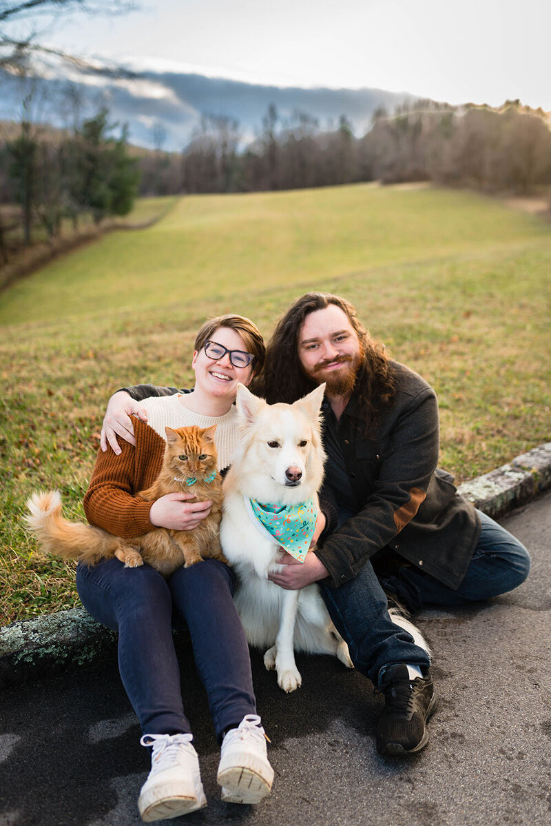 A Virginia wedding photographer sits on a curb with her  husband, dog, and cat during a ten-year anniversary session  at an overlook along the Blue Ridge Parkway.