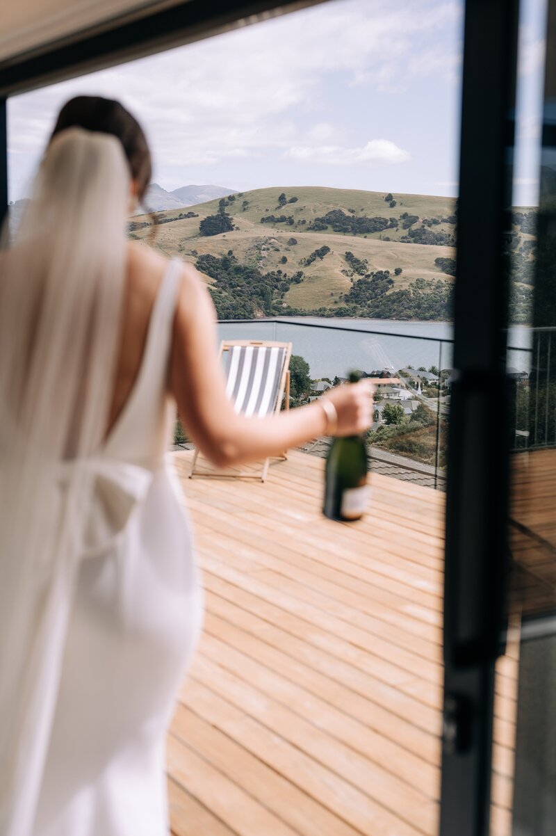 bride with champagne bottle walks across airbnb balcony in akaroa on wedding morning