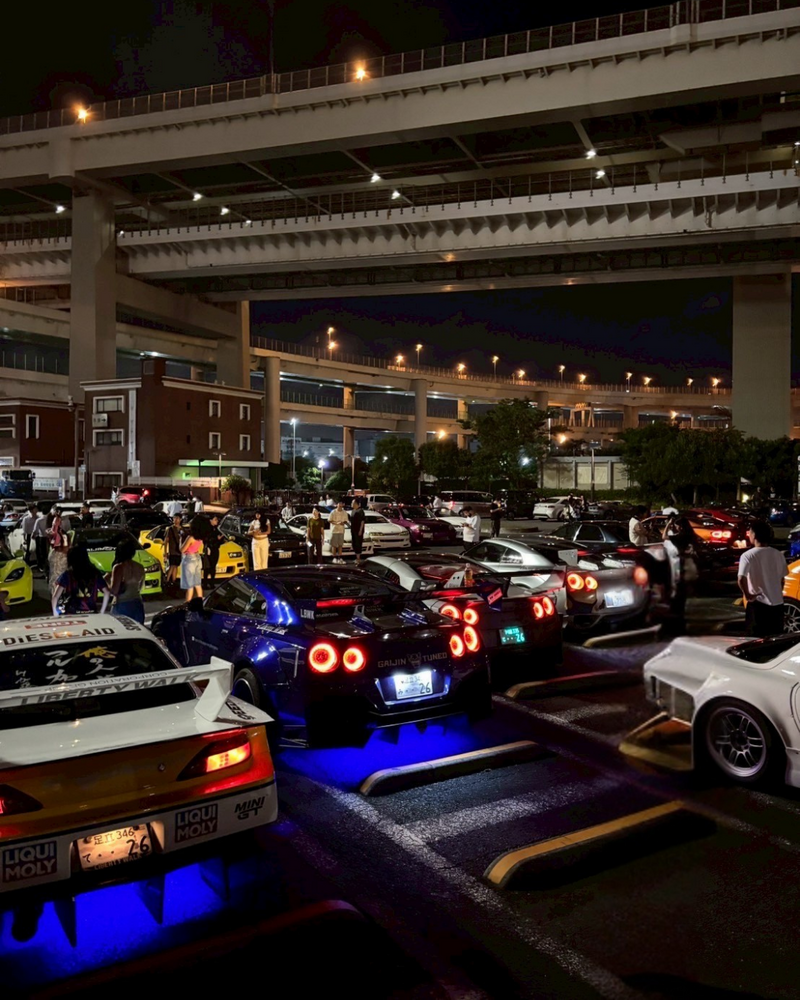 A glossy black Ferrari California parked under the illuminated highway overpasses at Daikoku Parking Area in Japan, surrounded by other cars and nighttime city lights.