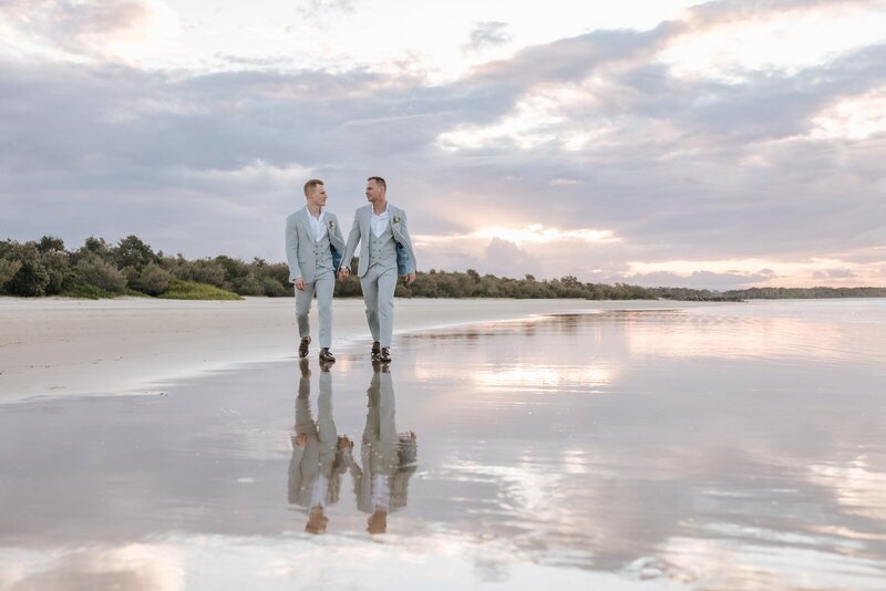 Two men on their wedding day walking along the beach holding hands