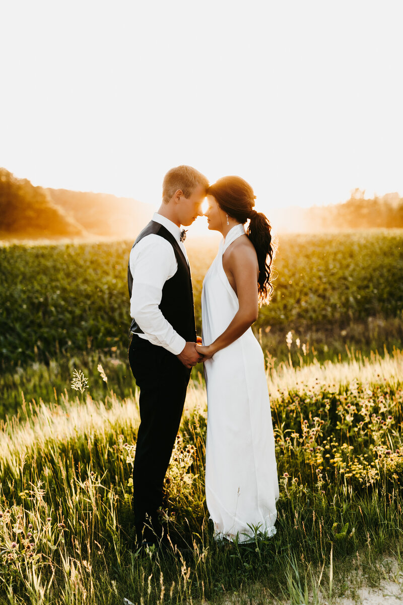 Bride and groom holding hands with their heads together in a field with the sun setting in the background