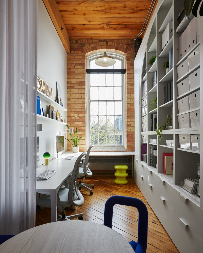 A tall, narrow room with warm brick and wood accents, modern white desks, office chairs, and shelves, and a large paned window.