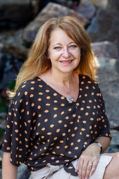 Cyndi Stewart sits on a rock outdoors, smiling warmly in a patterned blouse with her hair down.