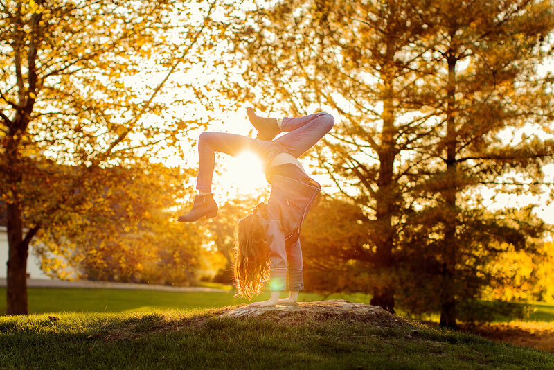 Tween girl is in jeans, long red, curly hair, doing a gymnastic pose with the sun setting behind her.
