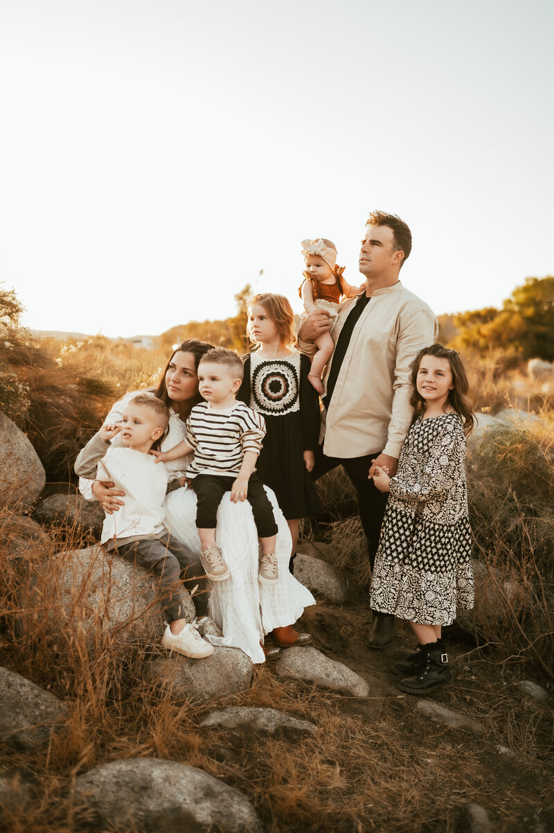 Family of six wearing coordinating outfits all face the beautiful sunset.