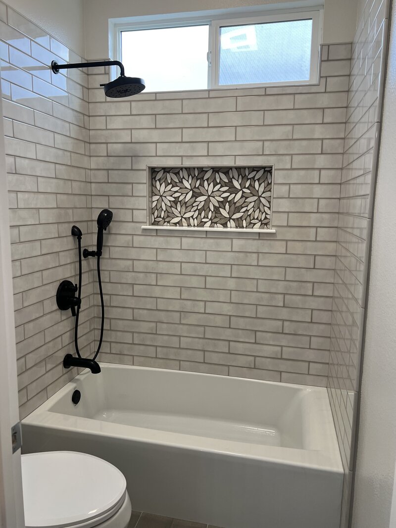 A tub shower combo with grey subway tiles and a decorative tiled shampoo box and black hardware.