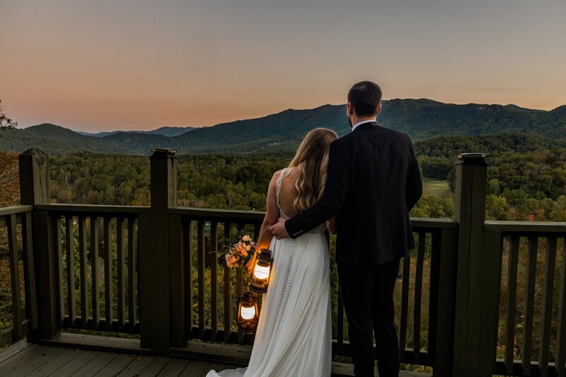 A bride and groom stand looking out at the sun set behind a vast mountain range after eloping. The groom has his arm around the bride while she leans her head on him. The sky is pink, blue and orange from the sunset while the vast mountain range is a mix of blue and green. They are both in wedding gear holding lanterns.
