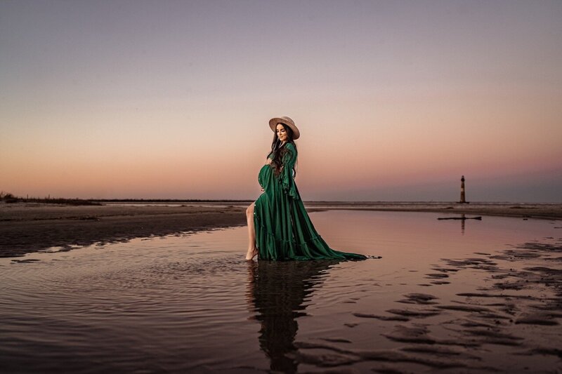 Pregnant woman in a flowing green dress walking on the beach at sunset during a maternity session