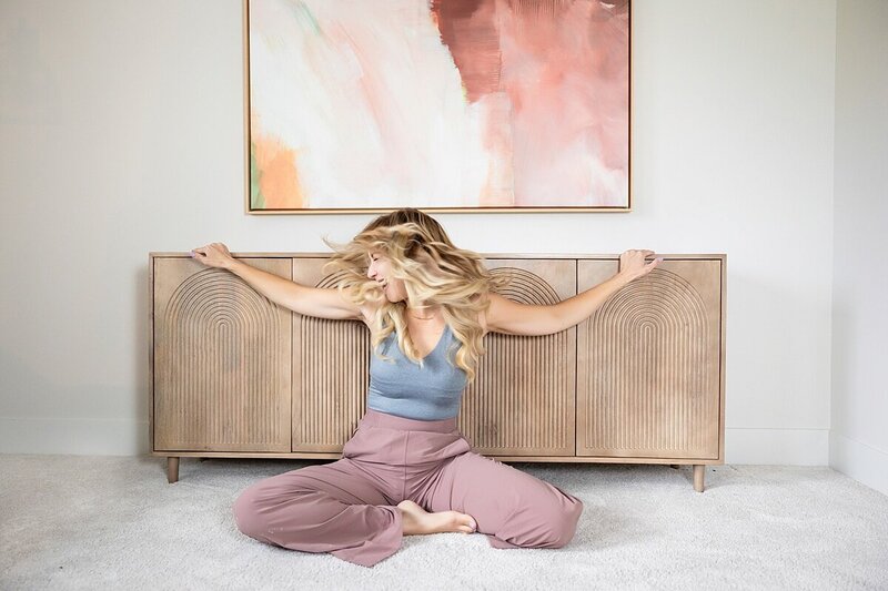Blonde woman sits cross-legged on the floor, tossing her hair playfully in front of a natural wood credenza and abstract artwork. Captured in a modern, light-filled space during a personal branding photoshoot, this image conveys creativity, freedom, and bold feminine energy.