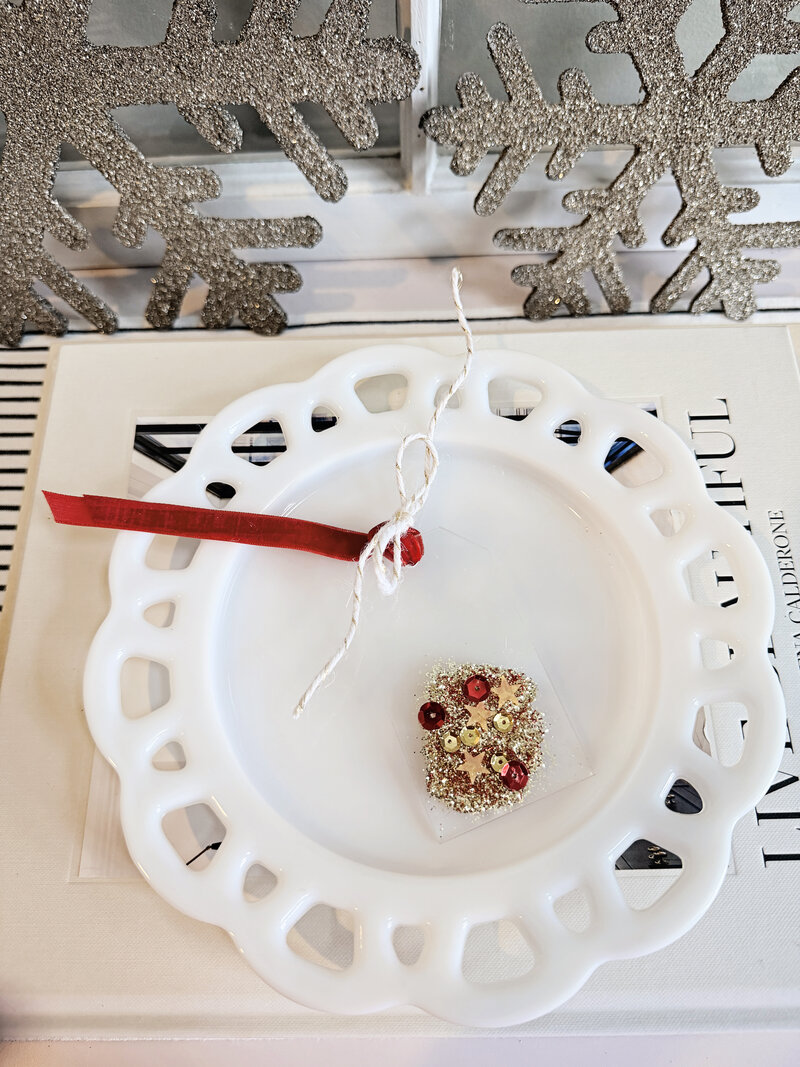 Mini acrylic Christmas gift tag from Sweet Pea Ink Creative embellished with gold glitter, red sequins, and gold stars, finished with red velvet ribbon. Displayed on white scalloped plate.