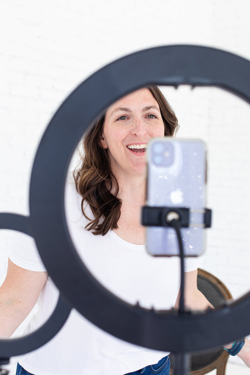 social media strategist is recording a reel on her iPhone. the cell phone is on a black tripod with a light and she is smiling in a white shirt and smiling at the camera