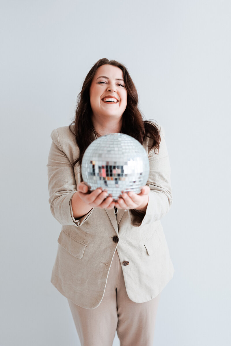 This is a image of Mariah of Moonlight Brand Design holding a disco ball and laughing. She is wearing a tan suit. 